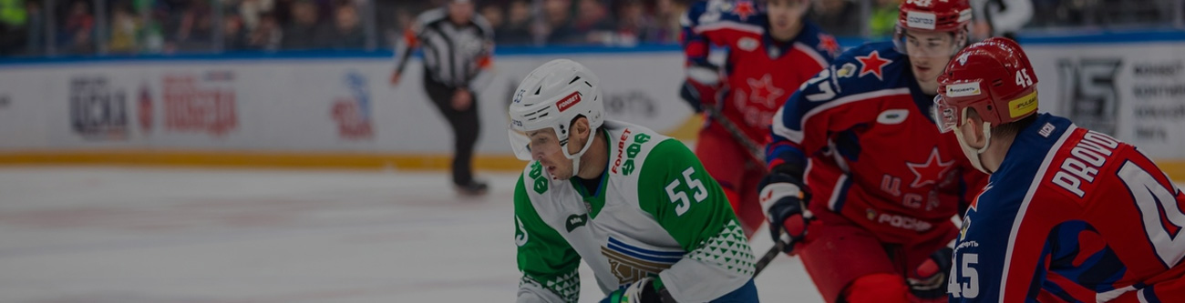 Игроки в хоккейной форме борются за шайбу на льду. Один в бело-зеленой форме с клюшкой, двое в красно-синей экипировке. На заднем плане судья и трибуны с болельщиками