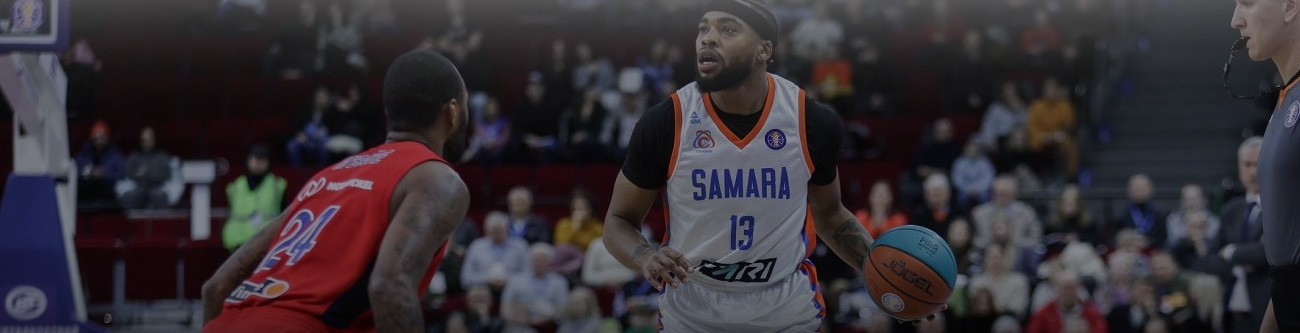 На изображении два баскетболиста в момент игры. Игрок в белой форме с мячом, другой в красной форме защищается. Вокруг видна публика на трибунах