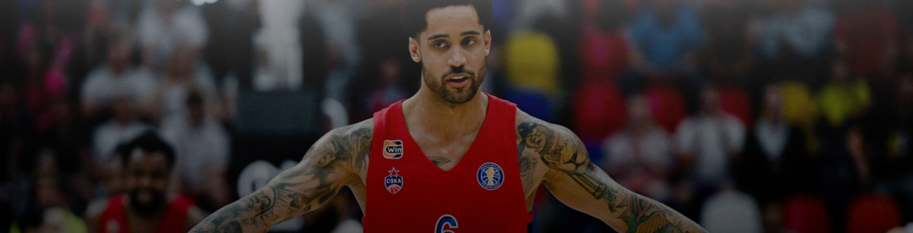 На изображении баскетболист в красной форме находится на площадке. Вокруг него размытая толпа зрителей. У спортсмена татуировки на руках и тёмные волосы