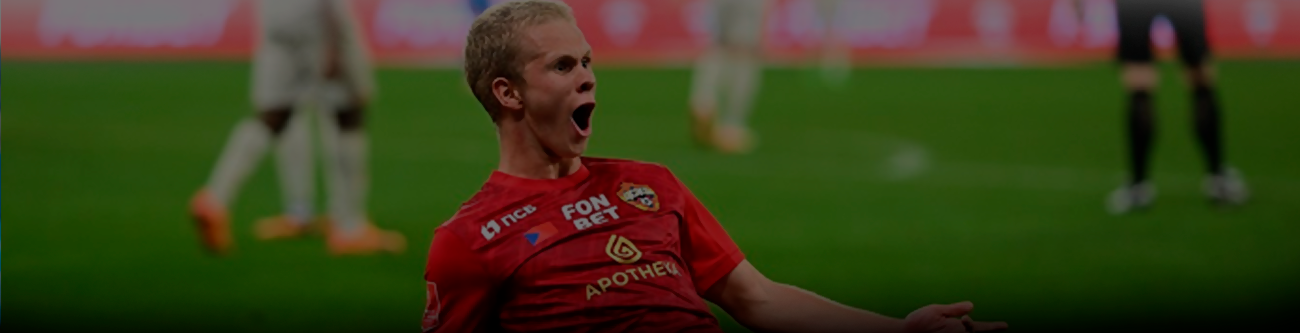 Футболист в красной форме с логотипом и надписью PFC празднует гол, скользя на коленях по траве. На заднем плане размытые фигуры игроков и красно-белые трибуны стадиона